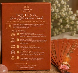 A brown cardboard box for gratitude cards with printed instructions titled 'HOW TO USE Your Affirmation Cards,' shown alongside a small fan, a crystal ball, and dried flowers in a softly styled setting.